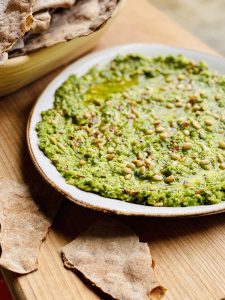 A bowl of brassica pesto which is a green coloured dip sprinkled with toasted pine nuts on top. It's seved in a rustic white bowel with a brown edge.